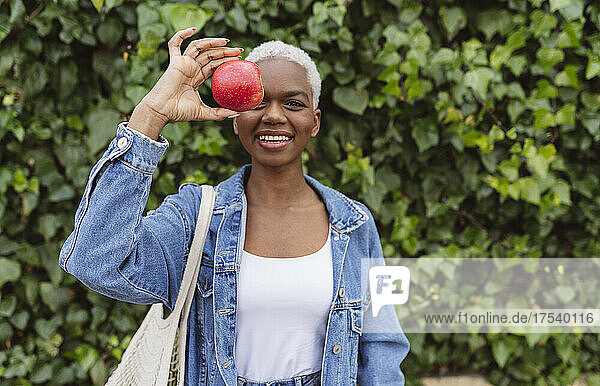 Woman covering eye with apple standing in front of plant