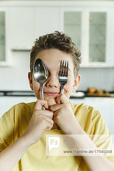 Smiling boy covering eyes with fork and spoon at home