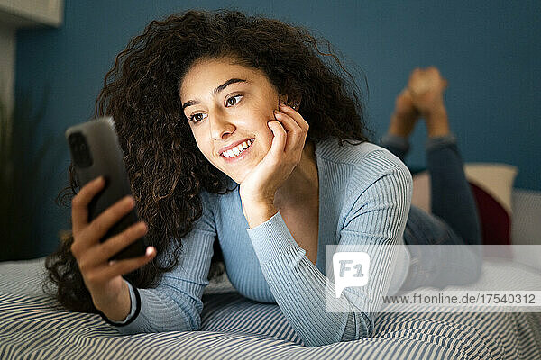 Woman with hand on chin using smart phone lying on bed