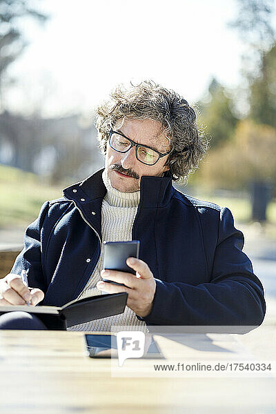Mann mit Brille schreibt an sonnigem Tag im Straßencafé mit dem Handy Tagebuch