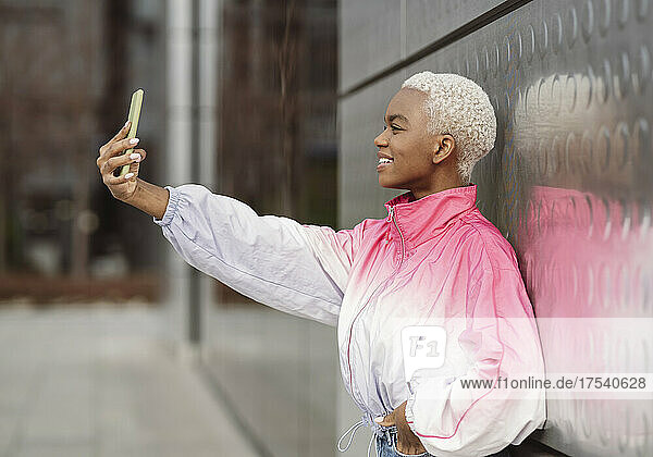 Young woman taking selfie through smart phone in front of wall