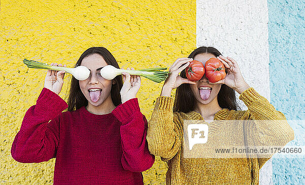 Playful sisters covering eyes with vegetables in front of multi colored wall