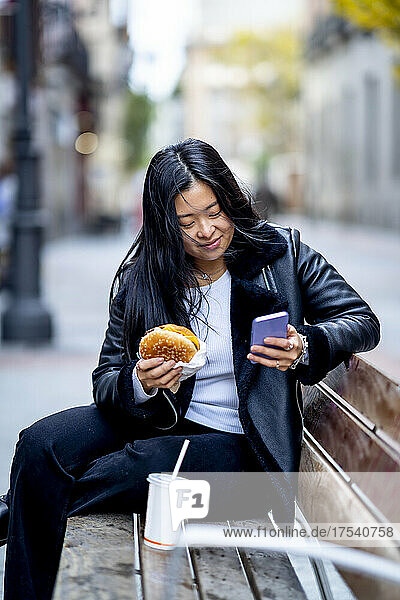 Frau mit Essen zum Mitnehmen per Handy auf Bank