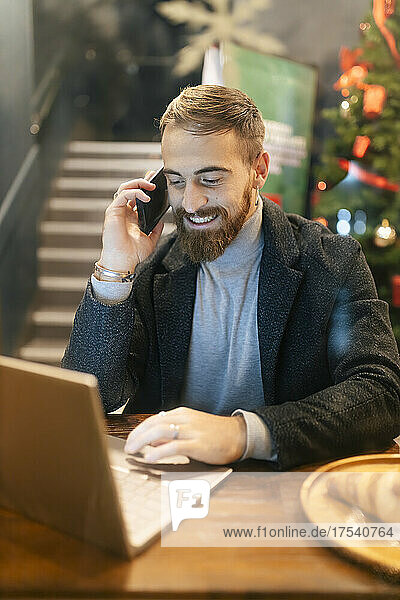 Lächelnder Geschäftsmann  der im Restaurant mit dem Mobiltelefon spricht und einen Laptop benutzt