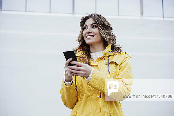Smiling young woman holding mobile phone in front of wall