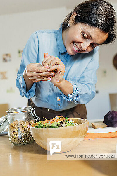 Smiling woman adding crushed walnuts in salad bowl on table at home