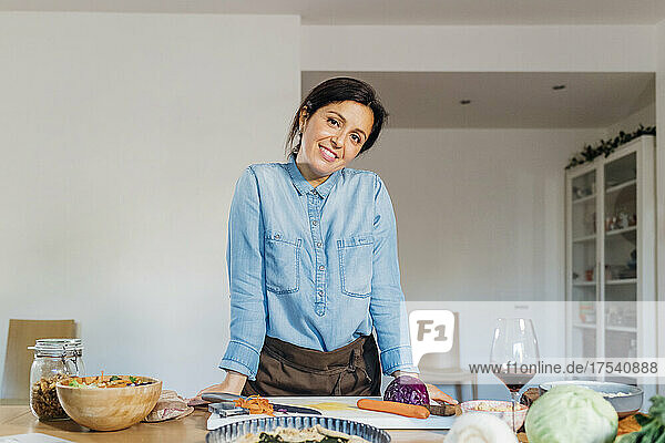 Smiling woman leaning on table with food and drink at home kitchen