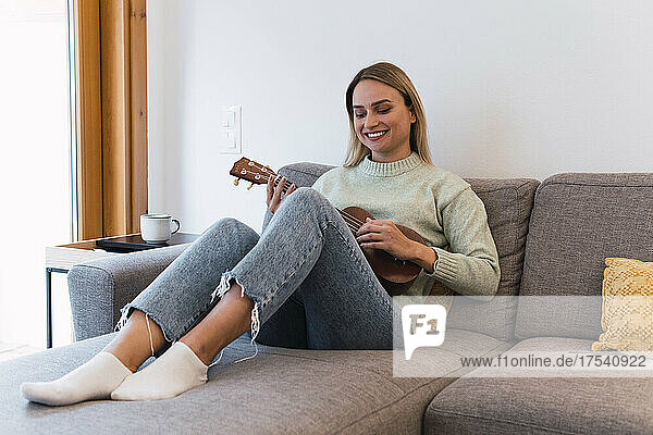 Lächelnde Frau spielt Ukulele und sitzt zu Hause auf dem Sofa