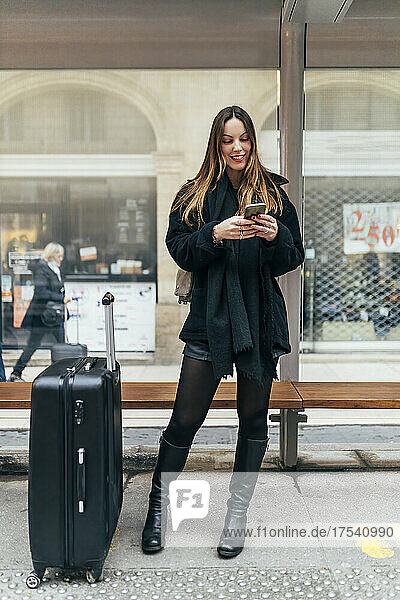 Smiling young woman with luggage using smart phone at tram station