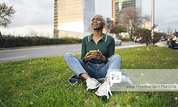 Smiling woman with smart phone sitting on grass in city