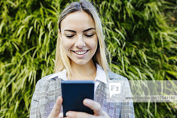 Smiling young businesswoman using mobile phone in front of green plants