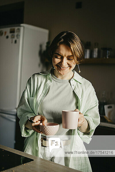Smiling woman holding coffee cup and bowl in kitchen at home