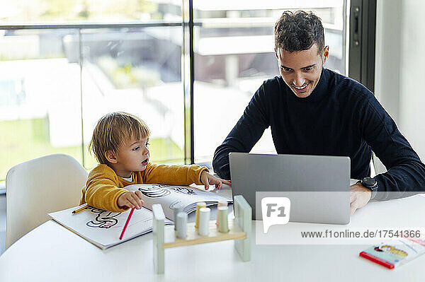 Smiling businessman freelancing on laptop by son at home