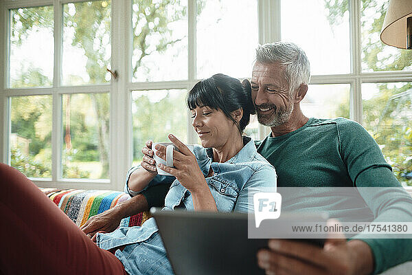 Happy man sharing tablet PC with woman holding coffee cup at home