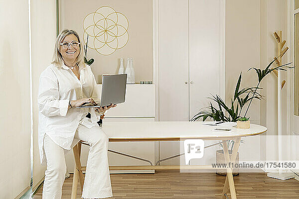 Smiling businesswoman holding laptop on desk at workplace