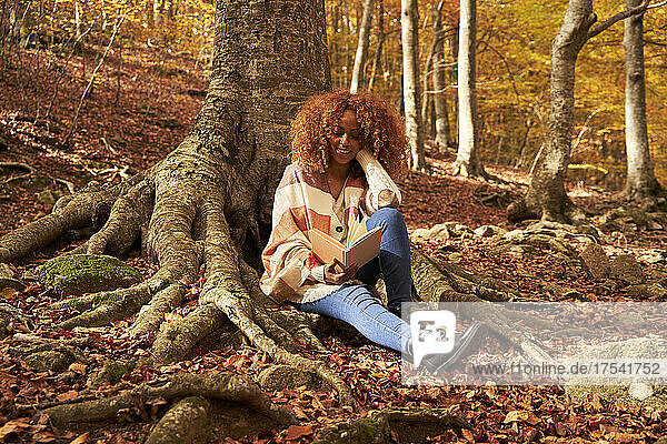 Young woman reading book in by tree in autumn forest