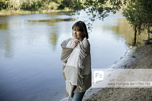 Thoughtful woman wrapped in blanket standing at riverbank