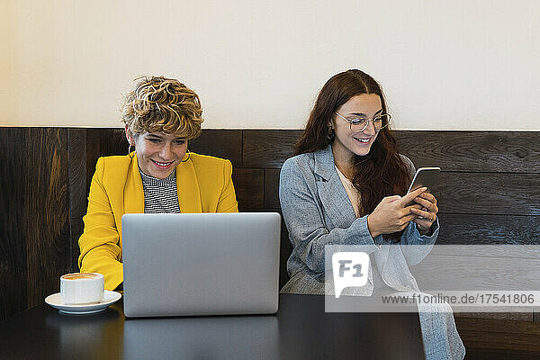 Lächelnde Geschäftsfrau  die von einem Kollegen am Laptop arbeitet und im Café ein Smartphone nutzt