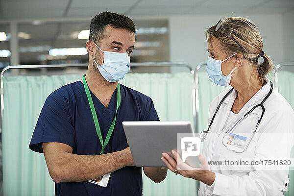 Krankenpfleger mit schützender Gesichtsmaske schaut den Arzt an  der im Krankenzimmer einen Tablet-PC hält