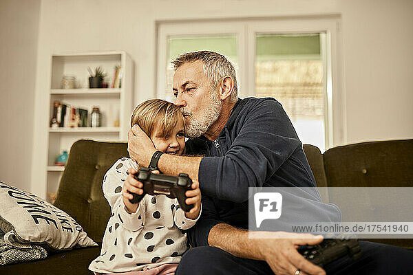 Grandfather kissing granddaughter holding joystick at home