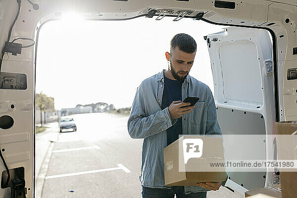 Delivery man using mobile phone holding box at van doorway