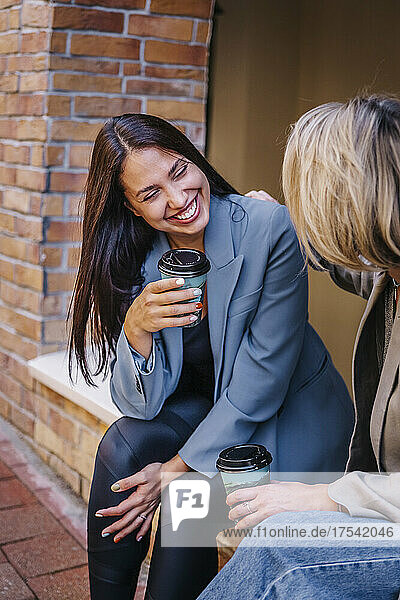 Smiling woman holding disposable cup talking with friend at wall