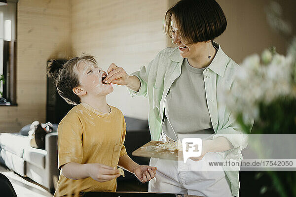 Lächelnde Mutter füttert ihren mit Mehl bedeckten Sohn zu Hause mit geriebenem Käse
