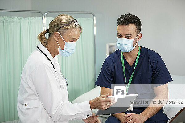 Ärztin mit schützender Gesichtsmaske diskutiert mit Krankenschwester über Tablet-PC im Krankenzimmer