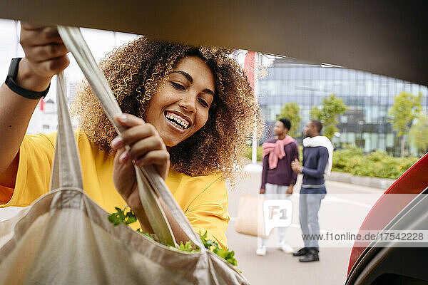 Lächelnde Frau lädt Einkaufstasche in den Kofferraum eines Autos und Freunde stehen im Hintergrund