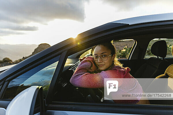 Woman leaning on steering wheel in car at sunset