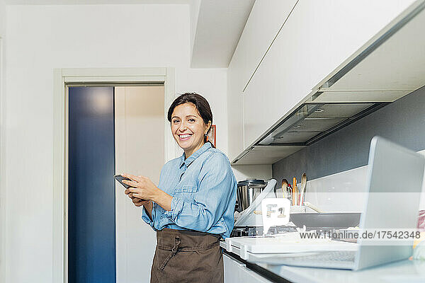Lächelnde Frau benutzt ihr Mobiltelefon und lehnt sich zu Hause an die Küchentheke
