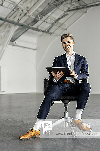 Smiling businessman holding tablet PC on chair at industial hall
