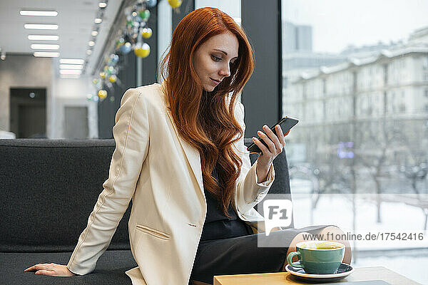 Businesswoman using smart phone sitting on sofa in office cafeteria