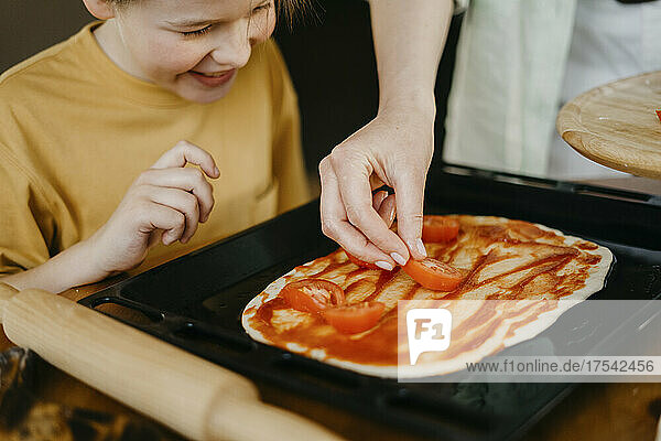 Junge schaut Mutter zu  die Tomatenscheiben auf den Teig im Tablett legt