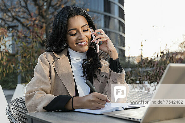 Lächelnde Geschäftsfrau  die Tagebuch schreibt und auf dem Smartphone spricht