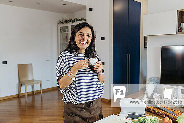 Smiling woman holding coffee cup standing by table at home
