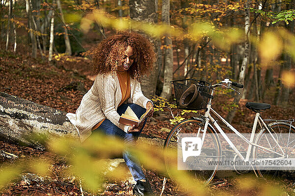 Woman sitting on log by bicycle reading book in autumn forest