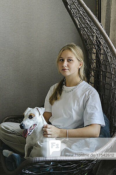 Teenage girl sitting with pet dog on hanging chair at home