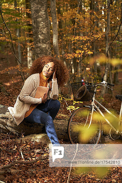 Woman with hand in hair holding book sitting on log by bicycle in autumn forest