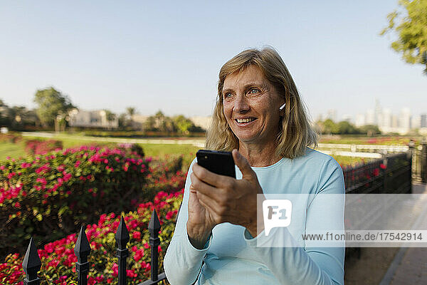 Lächelnde Frau mit In-Ear-Kopfhörern hält Smartphone am Zaun im Park