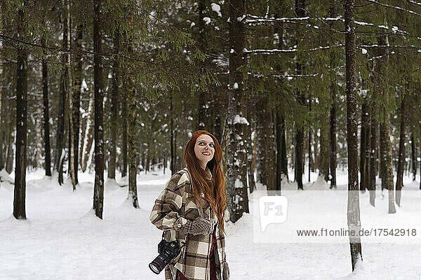 Smiling redhead woman with camera day dreaming in winter forest
