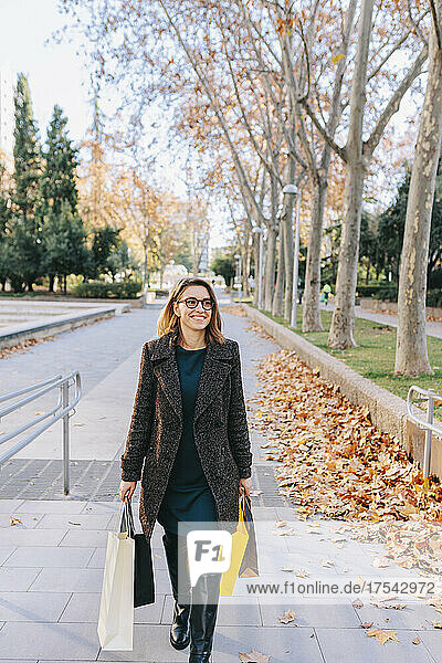 Smiling woman with shopping bags walking on footpath in autumn