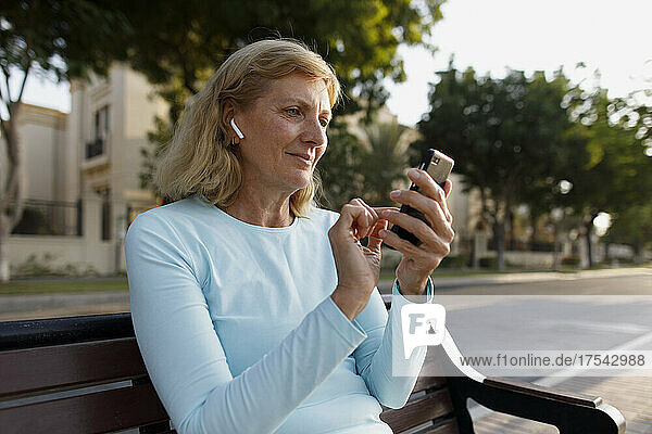 Ältere Frau mit In-Ear-Kopfhörern und Smartphone auf Bank sitzend