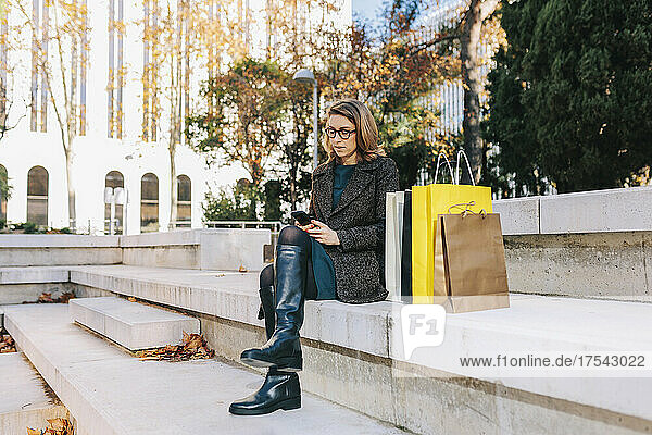 Woman using smart phone sitting on steps with shopping bags