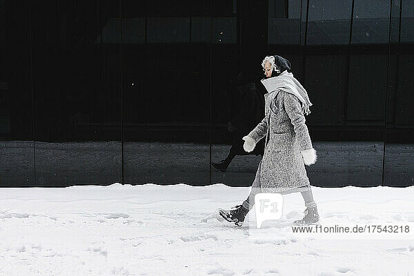 Woman wearing gray overcoat walking on snowy footpath by black wall