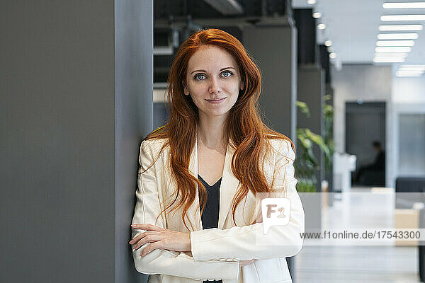 Redhead working woman with arms crossed leaning on column in office corridor