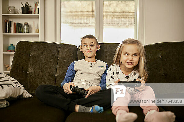 Brother and sister playing video game sitting on sofa at home