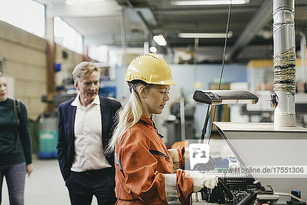 Weiblicher Industriearbeiter mit Schutzhelm bei der Arbeit an Maschinen  während im Hintergrund Manager in einer Fabrik zu sehen sind