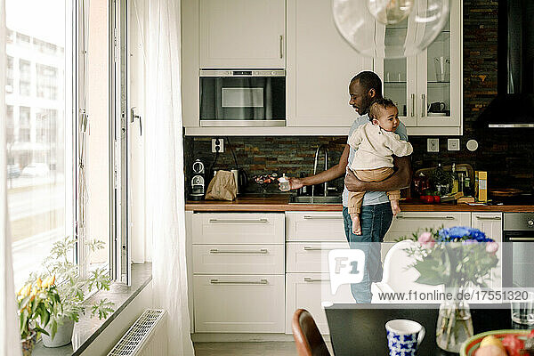Vater mit Babyflasche trägt männliches Kleinkind in der Küche zu Hause