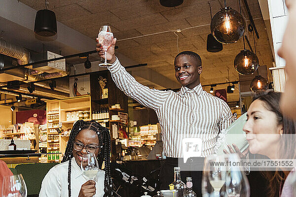 Lächelnder Mann mit Weinglas in der Hand zwischen männlichen und weiblichen Freunden während einer Dinnerparty in einer Bar